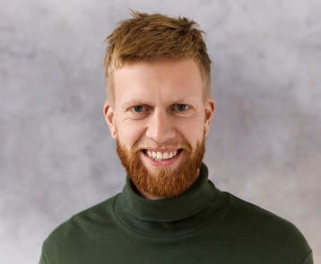 Portret van een lachende jongeman met rood haar en een rode baard, gekleed in een groene coltrui, gefotografeerd tegen een lichte, gestructureerde achtergrond.