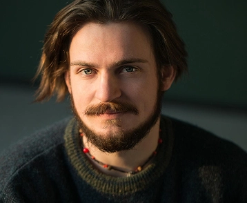 Portret van een jonge man met halflang haar en een baard, gekleed in een casual trui, gefotografeerd bij zacht natuurlijk licht.