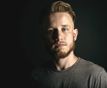 Portret van een jonge man met kort haar en een baard, gekleed in een grijs shirt, gefotografeerd tegen een donkere achtergrond met zachte studioverlichting.