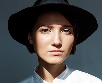 Portret van een jonge vrouw met een zwarte hoed en een witte blouse, die recht in de camera kijkt, met zachte belichting en een neutrale achtergrond.