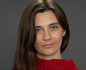 Portret van een jonge vrouw met lang bruin haar, gekleed in een rode trui, gefotografeerd tegen een neutrale achtergrond met een natuurlijke uitdrukking.